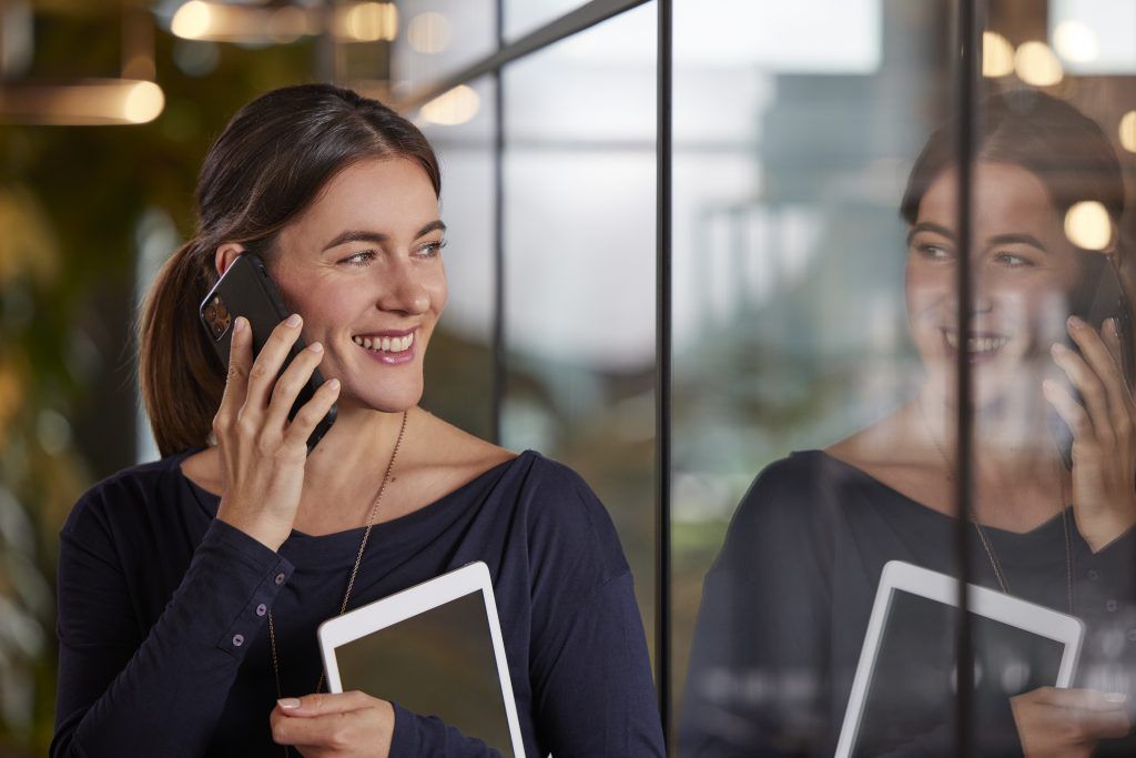 Frau hält Handy vor das Ohr und telefoniert. Außerdem hält sie ein Tablet in der anderen Hand.